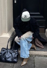 Schwarzer Mantel über einem dunklen Hoodie, helles, ausgeblichenes Denim und braune spitze Absätze. Schwarze Lederhandtasche neben der Person, die eine Mütze mit grünem Schirm trägt.