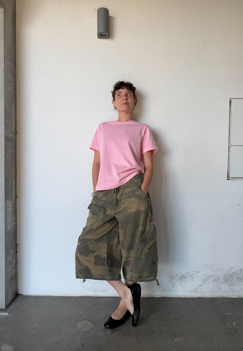 Personne se tient contre un mur blanc, portant un t-shirt rose, un pantalon cargo camouflage à jambes larges et des ballerines noires, les mains dans les poches et une jambe croisée.