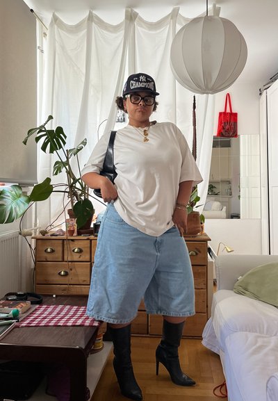 Camiseta blanca de gran tamaño, pantalones cortos de mezclilla azul claro y botas de tacón negro. Los accesorios incluyen una gorra negra y un collar de cadena dorada. Entorno interior.