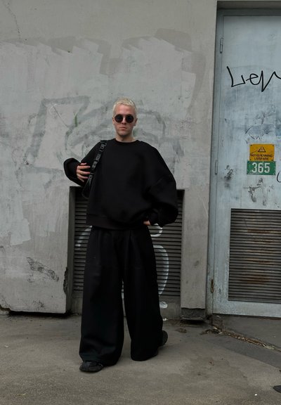 Sudadera negra de gran tamaño y pantalones de pierna ancha, combinados con gafas de sol oscuras y un bolso cruzado, frente a un fondo de graffiti.