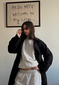 Manteau noir oversize, sweat-shirt gris et pantalon de survêtement assorti ; modèle portant des lunettes de soleil rectangulaires petites. Œuvre murale avec texte manuscrit.