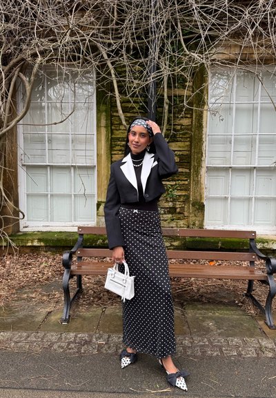 Mujer con falda de lunares en blanco y negro, blazer negro corto con solapas blancas, diadema a juego, sosteniendo un bolso blanco junto a un banco y una pared cubierta de enredaderas.