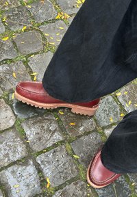 Red leather shoes featuring a rounded toe, raised stitching, and a prominent rubber sole, displayed against a cobblestone surface scattered with fallen leaves.