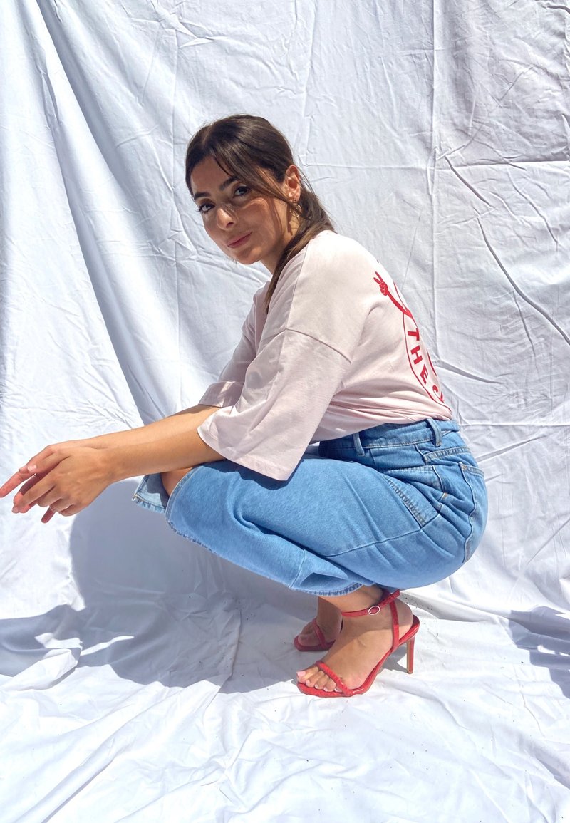 Világos rózsaszín, túlnyúló póló piros grafikával, kék, magas derekú farmernadrág és piros, pántos, magassarkú szandál, fehér háttér előtt.