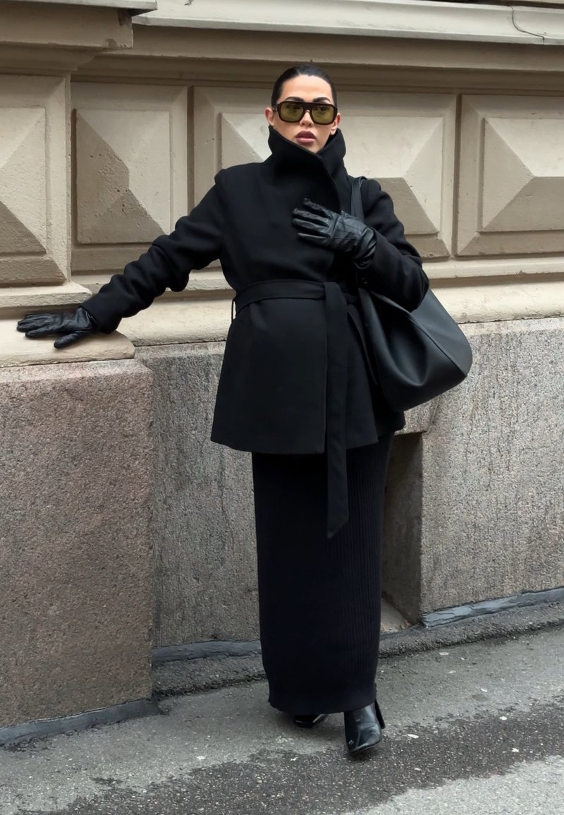 Femme en manteau noir, jupe longue, gants en cuir et lunettes de soleil, appuyée contre un mur en pierre, portant un grand sac à main noir à l'extérieur.