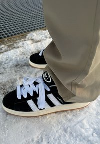 Fekete velúr sneaker fehér csíkokkal és fűzővel, gumitalppal és logós részlettel. Világos színű nadrág látható a cipő mellett.