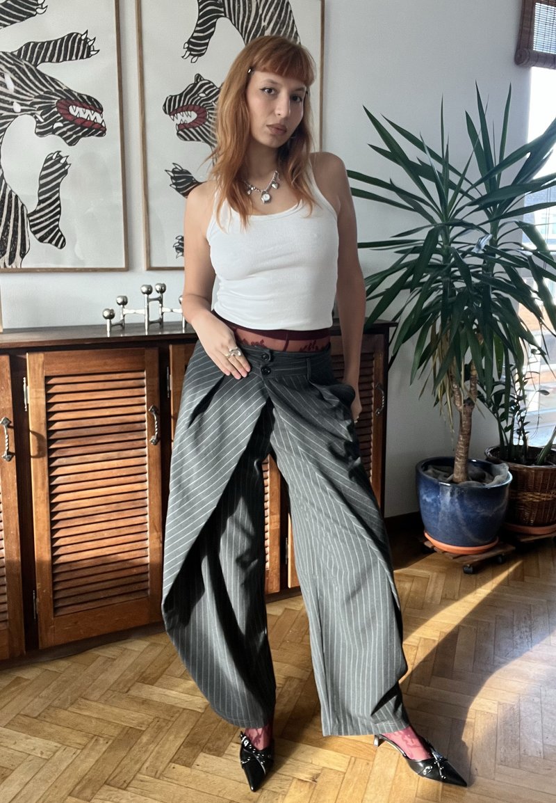 Γυναίκα με κόκκινα μαλλιά φοράει λευκή μπλούζα χωρίς μανίκια, φαρδιά παντελόνια με ρίγες, διάφανα ροζ καλσόν και μαύρα μυτερά παπούτσια, στέκεται σε δωμάτιο με ηλιακό φως.