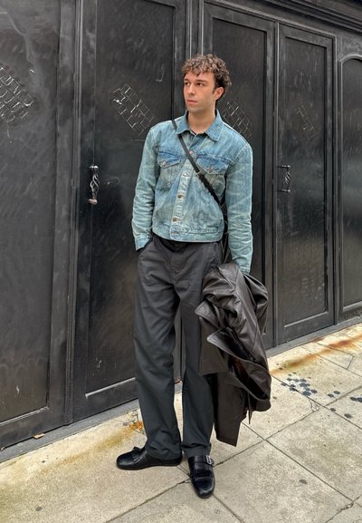 Joven con chaqueta vaquera, pantalones negros y zapatos sostiene una chaqueta de cuero oscura doblada mientras está de pie en la acera frente a unas puertas metálicas negras.