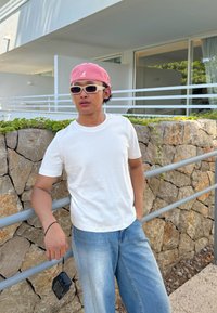 Witte korte mouwen t-shirt, lichte denim jeans, roze handdoekpet en zonnebril. Staande bij een textuursteen muur met groenvoorziening.