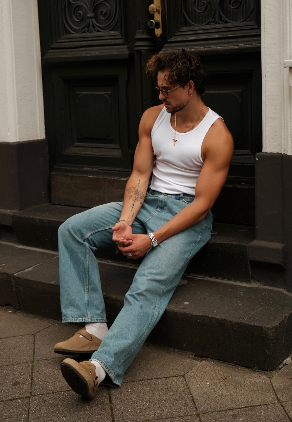 Top de tirantes blanco, jeans de mezclilla azul claro y zapatos beige. Sentado en escalones de piedra cerca de una puerta verde, mostrando un tatuaje visible en el brazo.