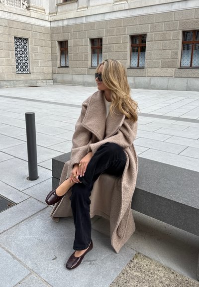 Abrigo oversize de mezcla de lana beige con un amplio cuello, combinado con pantalones negros texturizados y slippers marrones. Accesorios de joyería sencilla en las manos.