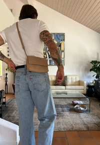 Beige crossover-veske laget av glatt skinn, rektangulær form, med klaffelukking, brukt over en hvit skjorte og paret med lyseblå jeans.