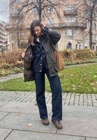 Olive zieleń, wodoodporna kurtka nałożona na denimowy ogrodniczki. Ciemnoniebieskie dżinsy o szerokich nogawkach, brązowe skórzane buty i brązowa torebka na ramię.