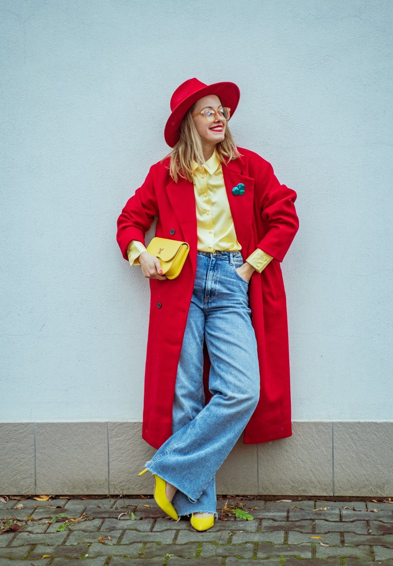 Kvinna lutande mot en vit vägg, klädd i en röd kappa och hatt, gul blus, gul väska, högklackade skor och ljusblå vida jeans.