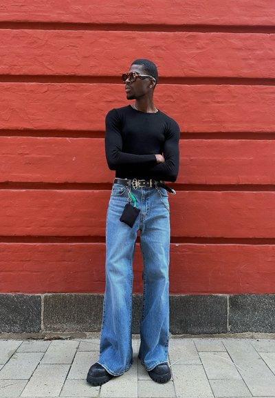 Top negro ajustado de manga larga, jeans azules de pierna ancha, zapatos negros gruesos y gafas de sol; accessorios con un cinturón y un pequeño bolso. Fondo de pared roja.
