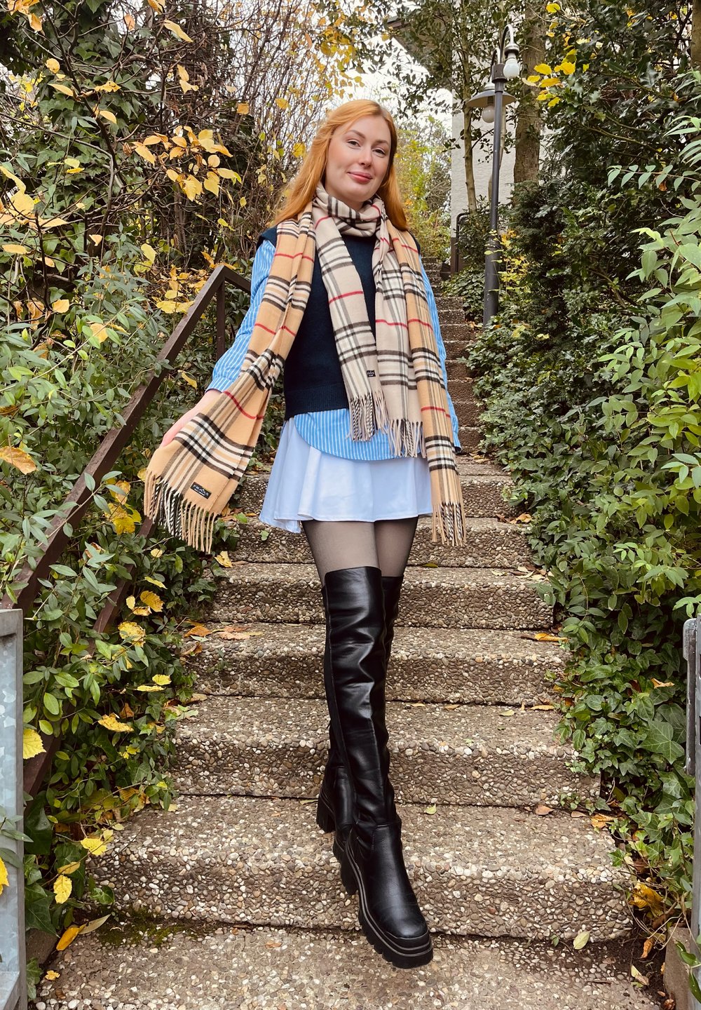 Bottes en cuir noires jusqu'aux genoux, écharpe beige à carreaux, chemise rayée bleue, jupe blanche à plusieurs couches, marches en pierre, verdure, feuilles d'automne.