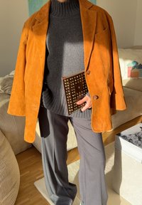 Rjavo rjavkasti žametni blazer, siv rebrast puli, široke sive hlače in teksturirana rjava torbica z zakovicami. Svetlo ozadje in pohištvo.