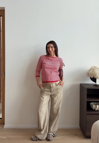 Mujer con camisa a rayas rojas y blancas y pantalones anchos beige, de pie con las manos en los bolsillos frente a una pared blanca lisa en el interior.