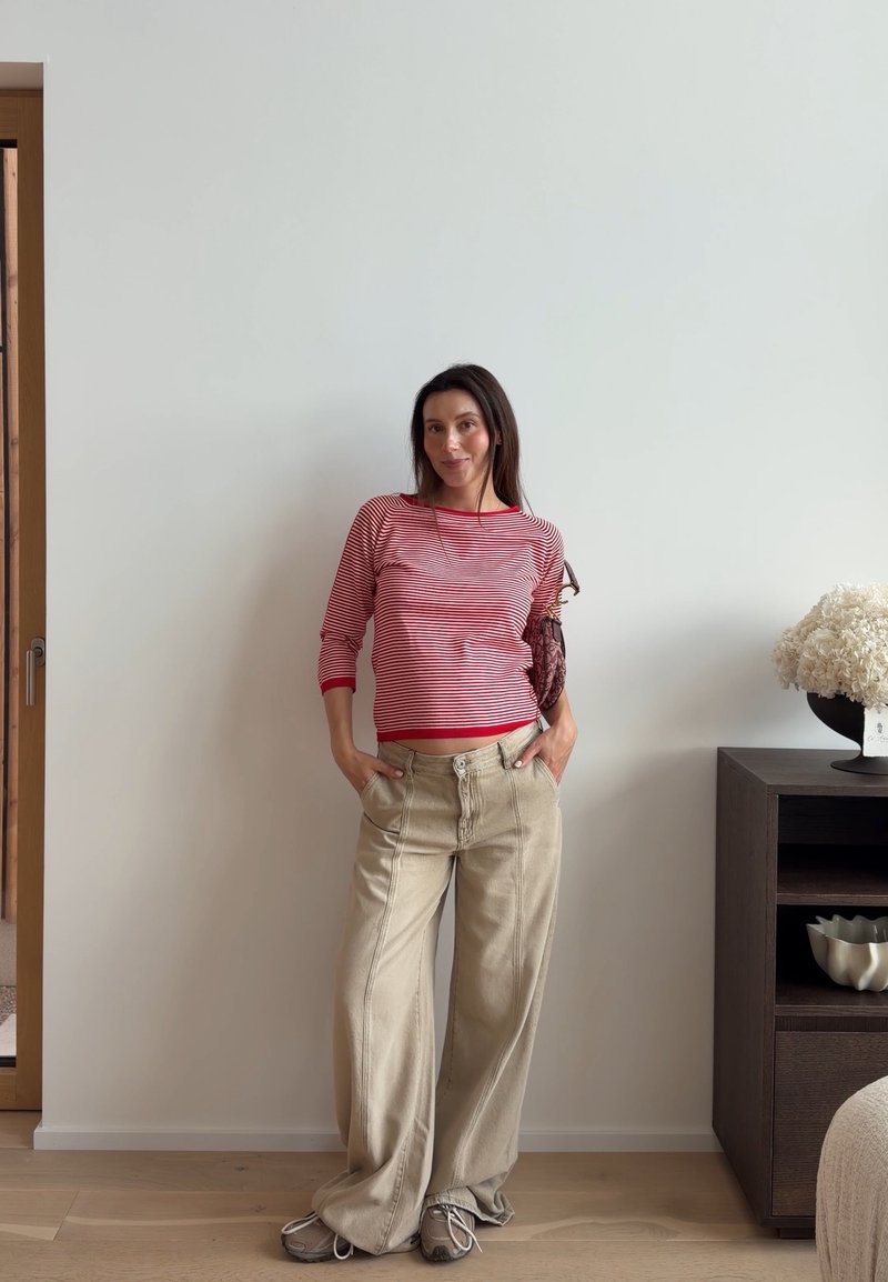 Femme portant une chemise rayée rouge et blanche et un pantalon large beige, debout les mains dans les poches contre un mur blanc uni à l'intérieur.