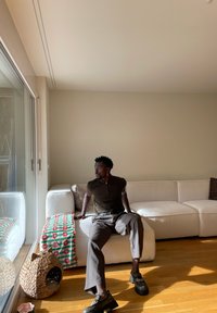 Muž v tmavé košili a hnědých kalhotách sedí na bílém rohovém gauči vedle zeleno-červeně pruhované deky v místnosti osvícené sluncem.