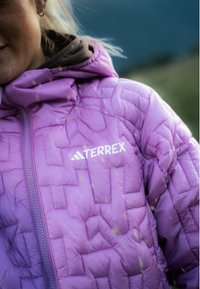 Giacca trapuntata viola con zip frontale e logo "TERREX". La texture è imbottita con un motivo geometrico. Finitura in tessuto liscia.