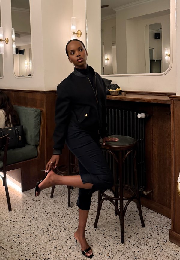 Una mujer vestida de negro posa junto a una mesa alta de cafetería, manteniéndose en equilibrio sobre un tacón en un restaurante con una iluminación cálida.