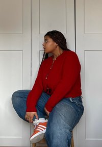 Κόκκινο πλεκτό καρντιγκάν, φαρδιά μπλε τζιν και πορτοκαλί αθλητικά με ρίγες. Πλεξούδα μαλλιών και στρωματοποιημένα κοσμήματα. Απλό φόντο με λευκές πόρτες.