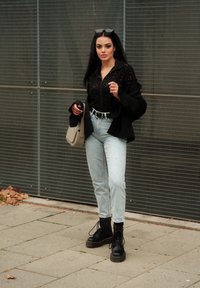 Μαύρο oversized σακάκι, μαύρες δερμάτινες μπότες με κορδόνια, ανοιχτό μπλε ψηλόμεσες τζιν, και μαύρο πουκάμισο με κουμπιά. Κρατώντας μια μπεζ τσάντα crossbody.