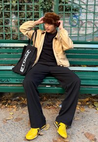 Veste en cuir beige, pull noir et pantalon noir. Chapeau en fourrure marron. Chaussures de sport jaunes avec des accents noirs. Sac fourre-tout noir avec un logo blanc.