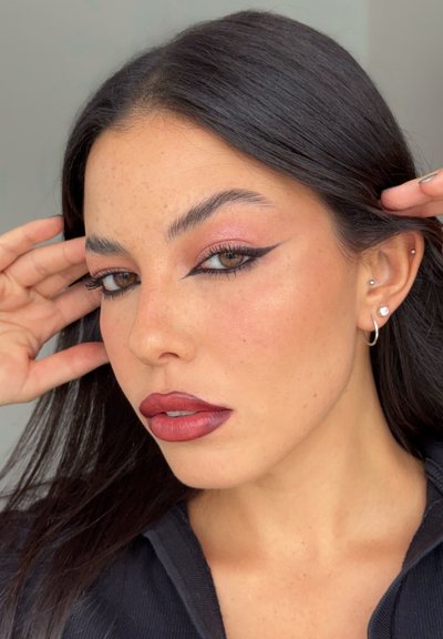 Mujer joven con cabello oscuro, delineador de ojos negro alado, sombra de ojos malva, labios rojos, pecas y múltiples perforaciones en las orejas, tocándose el cabello.