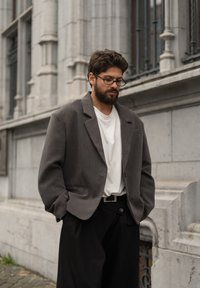 Šedé strukturované sako přes bílé tričko, doplněné černými kalhotami s širokými nohavicemi. Má knoflíky, přední kapsy a ležérní střih.