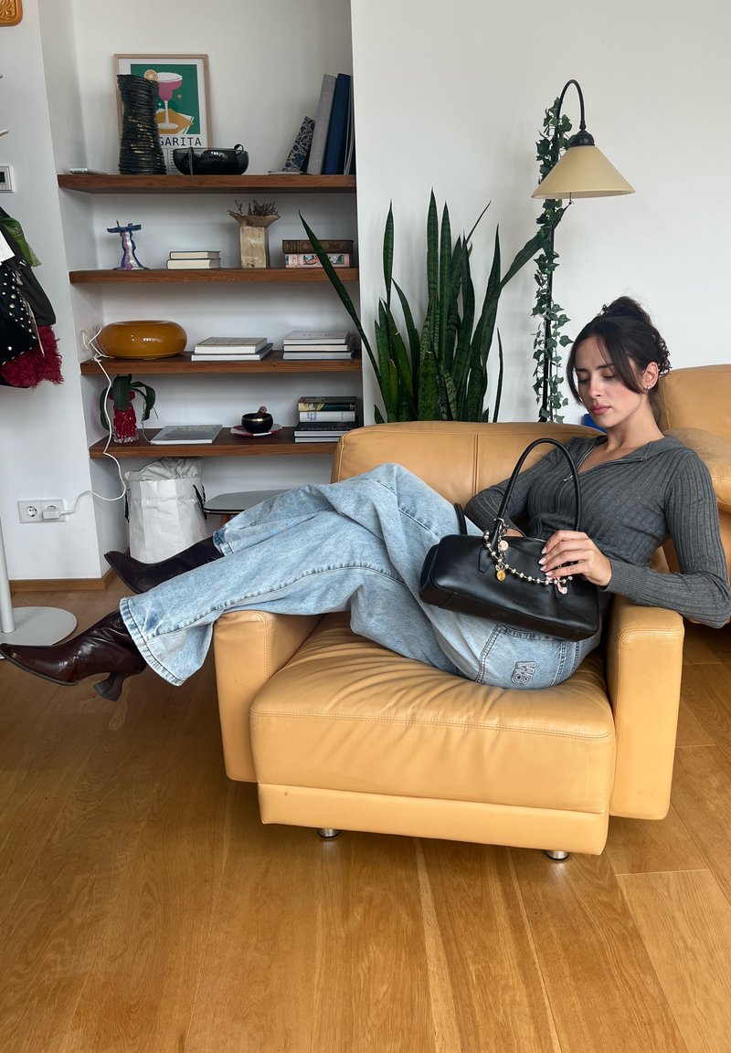 Bolsa preta com detalhe em corrente, suéter cinza canelado, jeans de perna larga azul-clara, botas de salto castanhas, sentada numa cadeira de braço em pele bege.