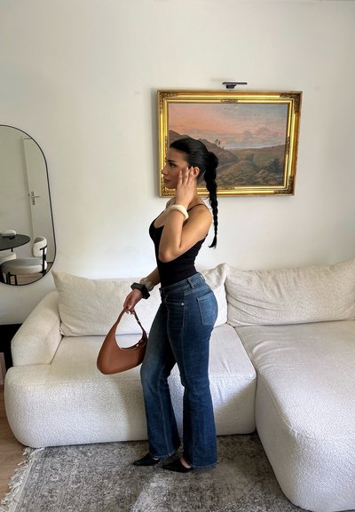 Mujer con el cabello largo y trenzado, vestida con top negro y vaqueros azules, está de pie junto a un sofá blanco sosteniendo un bolso marrón dentro de una sala de estar minimalista.