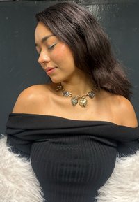 Svart off-shoulder ribbet topp med en gull- og sølvfarget hjerteanheng-kjede. Overflaten er glatt med en myk tekstur. Hvit faux pels aksenter.