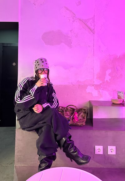 Chaqueta negra de gran tamaño con rayas blancas, pantalones negros holgados, botas negras hasta la rodilla, gorro gris estilo casco, sosteniendo una taza blanca en un espacio iluminado en rosa.