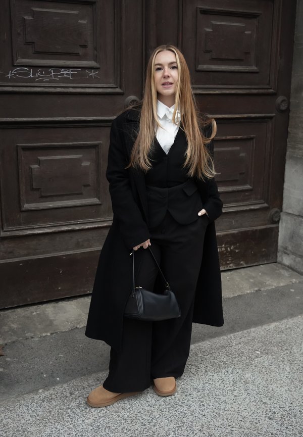 Femme aux longs cheveux blonds, portant un manteau noir, un pantalon large, une chemise blanche, des chaussures couleur beige, tenant un sac à main noir, debout devant des portes en bois.