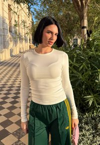 Femme aux cheveux courts noirs portant une chemise blanche à manches longues et un pantalon vert Adidas, tenant un sac rose, debout à côté de plantes sur un chemin extérieur carrelé.