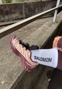 Stopalo resting na betonskom rubu nosi maroon i bež Salomon X Ultra 4 Gore-Tex planinarsku cipelu i belu čarapu sa crnim Salomon logom.
