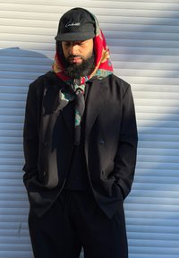 Giacca doppiopetto nera sopra una maglia nera, abbinata a pantaloni neri. L'outfit è completato da una sciarpa a fantasia multicolore e un berretto nero.