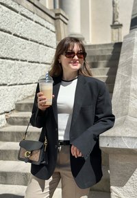 Musta ylisuureneva bleiseri, valkoinen vartalonmyötäinen paita, beige housut, kilpikonnakuosiset aurinkolasit, juoma kädessä ja musta laukku, jossa on kuvioitu pinta.