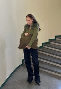 Olivově zelený svetr na zip s kožíškovým límcem, tmavé džínové zvonové kalhoty a hnědá kabelka ve tvaru polštáře s texturou, stojící na schodech.