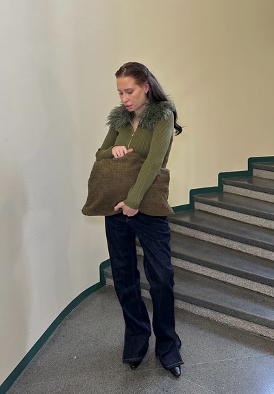 Suéter verde oliva con cremallera y cuello de piel, vaqueros acampanados de mezclilla oscura y un bolso marrón con textura en forma de almohada, de pie en las escaleras.