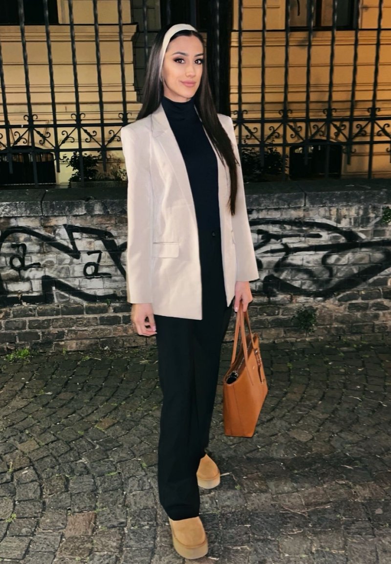 Femeie cu păr lung și închis la culoare, purtând un sacou bej, pantaloni negri și ținând o geantă maro, stă pe o stradă pietruită noaptea.