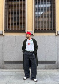 Άνδρας που φορά κόκκινο σκουφί, γυαλιά ηλίου, λευκό αθλητικό μπλουζάκι, μακρύ σκούρο παλτό, φαρδιά σκούρα παντελόνια και λευκά αθλητικά παπούτσια στέκεται με τα χέρια στις τσέπες.