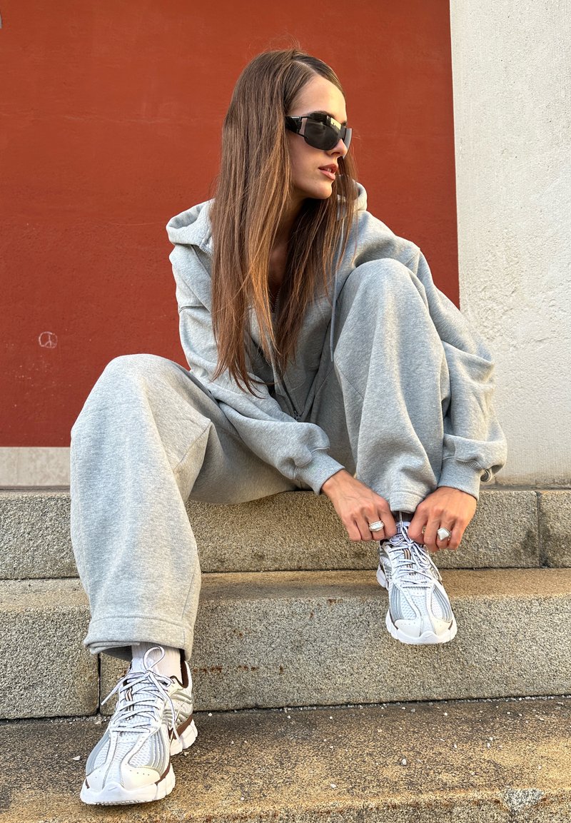 Ung kvinna med långt hår och solglasögon knyter vita sneakers medan hon sitter på stensteg mot röda och vita väggar.