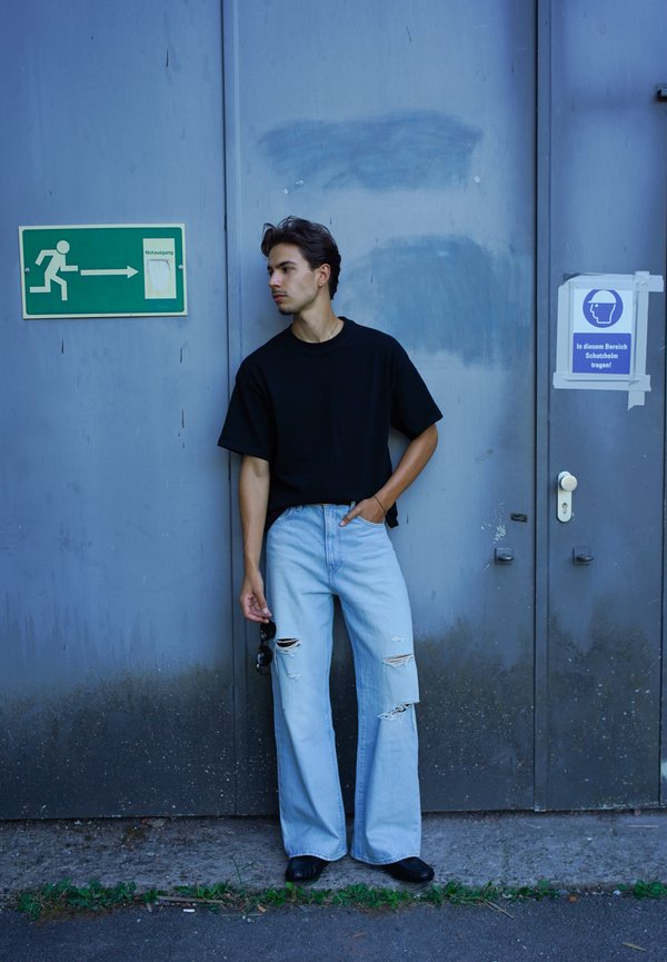 Jeune homme portant un t-shirt noir et un jean bleu clair déchiré, debout contre une porte métallique grise près d'une sortie de secours et de panneaux indiquant le port du casque.