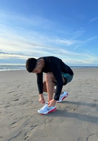 Baskets de sport blanches avec des accents rouges, tige texturée et logo Swoosh noir. Individu à genoux sur le sable, en train de lacer ses chaussures.