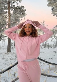 Jonge vrouw in een roze hoodie en joggingbroek die haar capuchon buiten in een besneeuwd bos aanpast tijdens een roze zonsondergang met lichtsnoeren aan de reling.