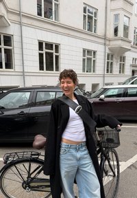 Czarny płaszcz, biały t-shirt, jasnoniebieskie dżinsy. Czarny rower z brązowym siodełkiem i przednim koszykiem, zaparkowany na ulicy z samochodami i budynkami.