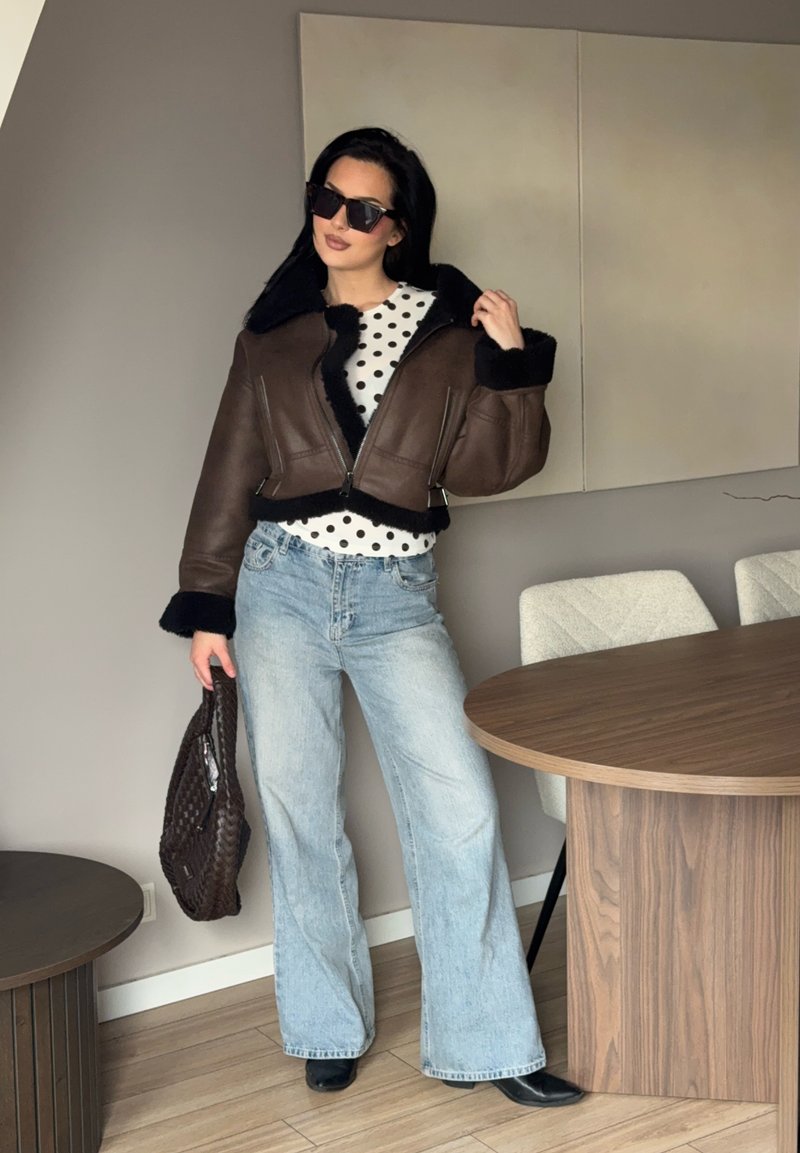 Femme portant une veste en peau lainée marron foncé, chemise blanche à pois, jean bleu clair évasé, bottes noires, lunettes de soleil, tenant un sac à main tressé marron à l'intérieur.